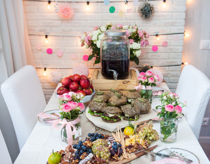 Decorated party table. Lemonade stand, apples, burgers, cheese and charcuterie platter with grapes, cheese, bread sticks, pecan nuts and canape. String ligts, flowers and paper balls decoration.