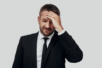 Frustrated young man in full suit suffering from headache while standing against grey background