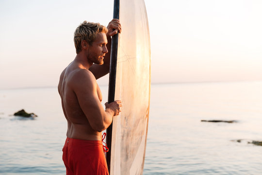 Image of young surfer man holding his surfboard by ocean at sunrise