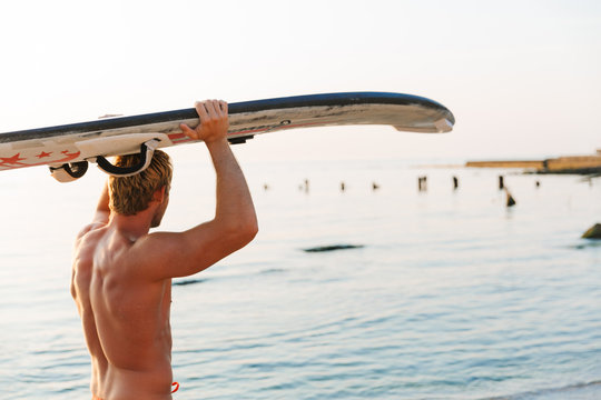 Image of caucasian surfer man carrying his surfboard by ocean at sunrise
