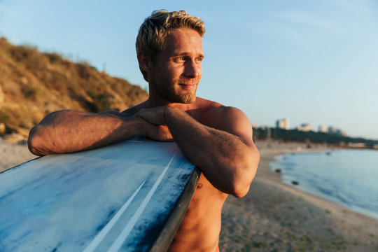 Image of sporty surfer man leaning on his surfboard by ocean at sunrise