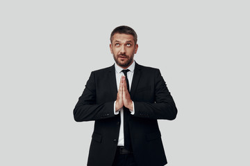 Charming young man in full suit keeping hands clasped while standing against grey background