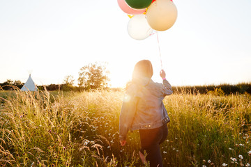 Rear View Of Woman Camping At Music Festival Running Through Field With Balloons Against Flaring Sun © Monkey Business