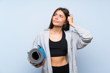 Young sport girl with mat over isolated blue background having doubts and with confuse face...