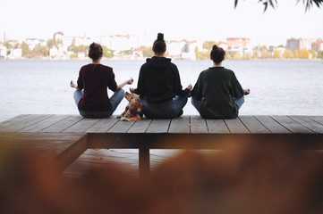  Three young women doing yoga at nature. Fitness, sport, yoga and healthy lifestyle concept - group of people making yoga pose on lake pier at sunset