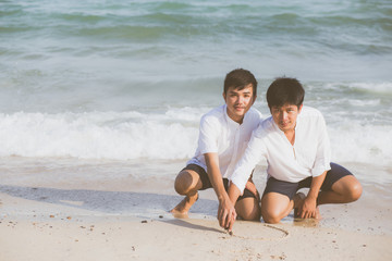 Fototapeta na wymiar Young asian couple gay smiling romantic drawing heart shape together on sand in vacation, homosexual happy and fun with love sitting on sand at the beach in travel summer, LGBT legal concept.