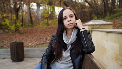 A young beautiful woman in casual clothes is sitting on a bench in autumn park.
