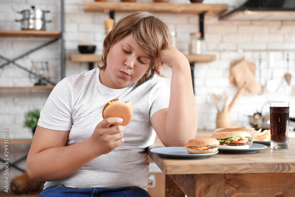 Poster Overweight boy at table with fast food in kitchen