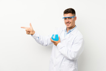 Young scientific holding laboratory flask over isolated background pointing finger to the side