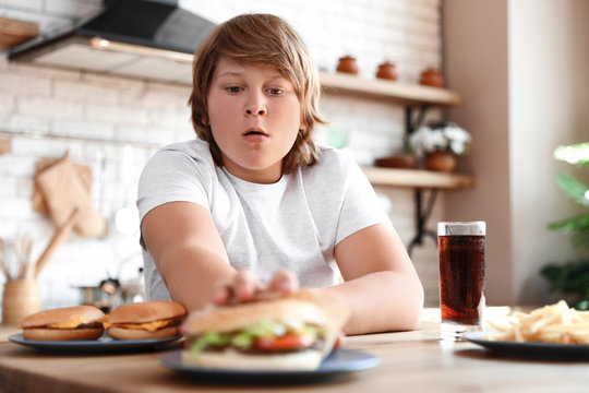 Emotional overweight boy at table with fast food in kitchen