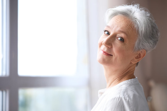 Happy mature woman near window indoors, space for text. Smart aging