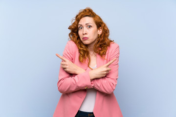 Redhead woman in suit over isolated blue wall pointing to the laterals having doubts