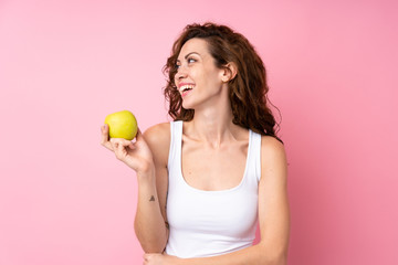 Young woman with curly hair holding an apple over isolated pink background