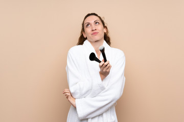 Young woman in a bathrobe holding makeup brushes making doubts gesture while lifting the shoulders