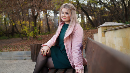 A young beautiful woman in casual clothes is sitting on a bench in autumn park.