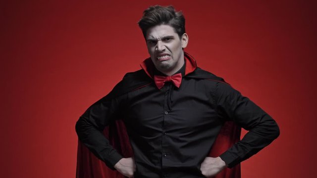 Surprised vampire man with blood and fangs in black halloween costume becoming unhappy and angry because of getting bad news isolated over red wall