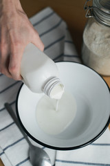 caucasian womans hand holds plastic bottle, pours kefir in white metal bowl on wooden table, cooking homemade oladyi, pancakes with wholegrain flour, top view of vertical life style stock photo image