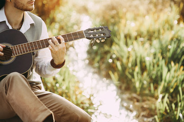 Young man playing guitar sitting on the bank of a mountain river on a background of rocks and...