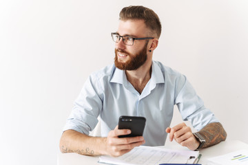 Image of happy young man holding smartphone while working in office