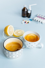 Home treatment versus medicaments. In the foreground, a cup of tea, a saucer of lemon and honey. In the background, various pills, a remedy for flu and runny nose. Loss of strength. Heatlhcare concept
