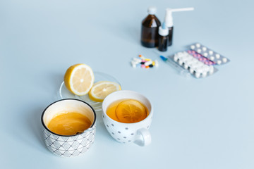 Home treatment versus medicaments. In the foreground, a cup of tea, a saucer of lemon and honey. In the background, various pills, a remedy for flu and runny nose. Loss of strength. Heatlhcare concept
