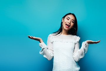 happy brunette asian woman gesturing on blue background