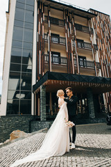 Stylish wedding couple standing near hotel. Young woman in white wedding dress and jewelry tiara with veil. Groom in a black suit.
