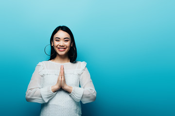 happy brunette asian woman with praying hands on blue background