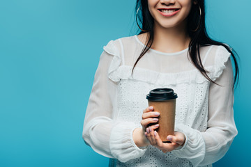 cropped view of smiling brunette asian woman holding coffee to go on blue background