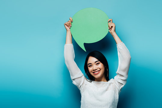 smiling brunette asian woman holding green speech bubble on blue background
