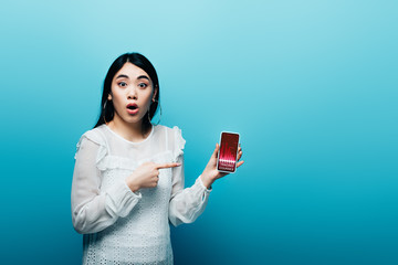 shocked asian woman pointing with finger at smartphone with trading courses app on blue background