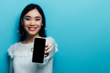 Naklejka premium selective focus of smiling asian woman in white blouse holding smartphone with blank screen on blue background