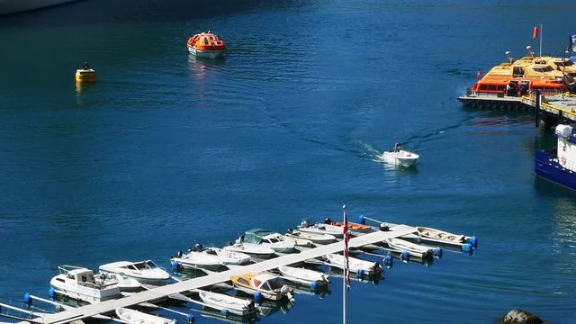 Aerial view of a marina with rows of motor boats and a small boat approaching a blue and white tour boat and orange and yellow ferry boats on calm blue water.