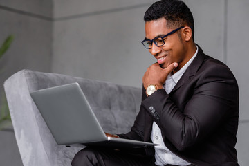 Portrait of a successful cheerful african-american businessman in formal suit working remotely with his international team using laptop and high speed internet while sitting in his office