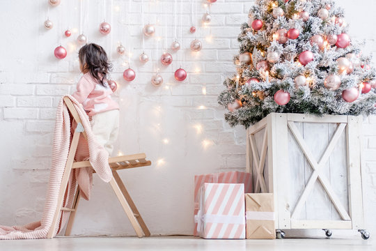 Little Curious Girl Stands On A Wooden Chair To Look Closer At The Christmas Toys Hanging As Decoration On The Wall Next To The Christmas Tree In A White Bright Interior Style. Xmas Home Decor Concept