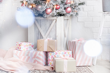 Christmas boxes with gifts decorated with wrapping paper and ribbons lie on light blurry background of white brick wall and artificial Christmas tree with pink toys in wooden box. Advertising space