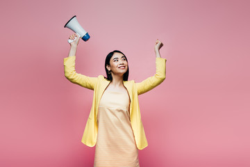 happy asian woman in yellow outfit holding megaphone isolated on pink