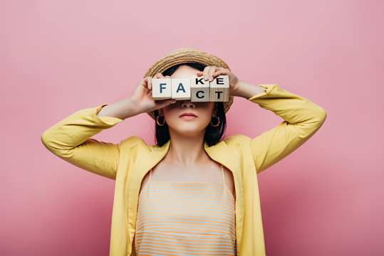 Asian Woman Holding Wooden Cubes In Front Of Face With Fake And Fact Lettering Isolated On Pink