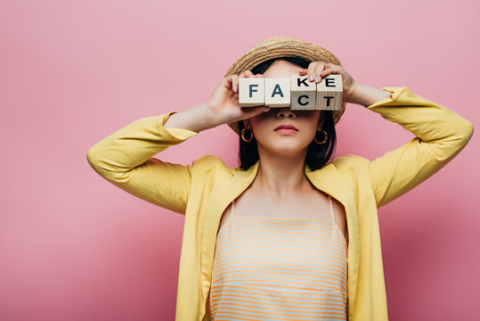 Asian Woman Holding Wooden Cubes In Front Of Face With Fake And Fact Lettering Isolated On Pink
