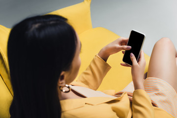 overhead view of asian woman holding smartphone while sitting on yellow couch isolated on grey