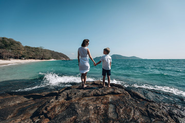 Loving mother and her little son hold hands and and look at the turquois sea standing on rocky beach