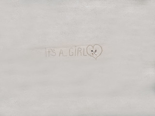 Future parents resting on the white sand of the beach by the turquoise waters enjoying anticipation, "It's a girl" written on the sand; drone view.