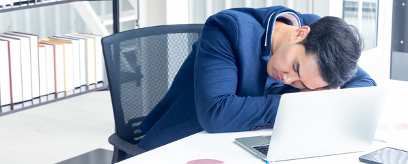 A young businessman sitting in a modern office. He has a feel sleepy because  hard work so tired weary fatigued and exhausted. On his table have a computer laptop tablet pen paper graph.