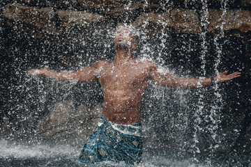 Fit and handsome topless Arabic man under the waterfall, huge rocks and water, splashes and flecks; natural beauty concept.