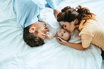 Happy mother kissing baby while lying on bed near smiling husband