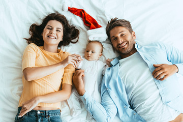 top view of happy parents lying on bedding near baby and santa hat