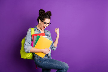 Profile side photo of delighted high school girl hold notebooks pass test achieve academic aims raise fists scream yeah wear denim jeans jacket green rucksack isolated shine violet color background