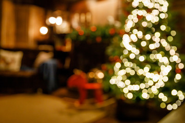Christmas and New Year interior - blur background: fireplace, lamps, green Christmas tree, brown leather sofa, gifts, candles, moose rocking chair. Lots of lights glowing in the dark.