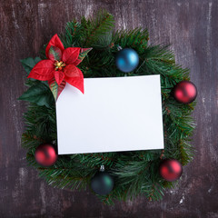 Fototapeta na wymiar Christmas wreath with decorations and blank white paper sheet on wooden background. New Year decoration. Top view, copy space.