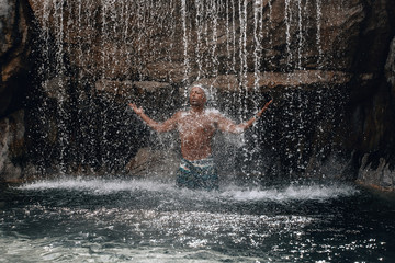 Fit and handsome topless Arabic man under the waterfall, huge rocks and water, splashes and flecks; natural beauty concept.